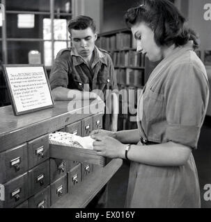 Historique, 1950, jeune femme bibliothécaire cherche dans un bac d'index de carte à la recherche d'un livre d'un jeune homme est à la recherche. Banque D'Images