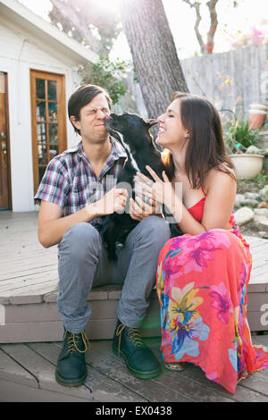 Jeune couple assis sur le patio avec dog licking face Banque D'Images