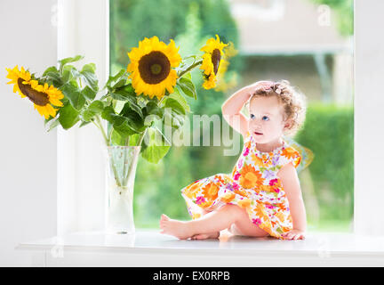 Happy smiling baby girl jouant avec de beaux tournesols sur une journée ensoleillée assis à une fenêtre pour le jardin Banque D'Images