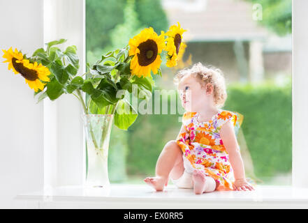 Happy smiling baby girl jouant avec de beaux tournesols sur une journée ensoleillée assis à une fenêtre pour le jardin Banque D'Images