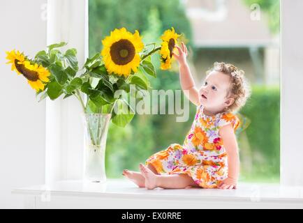 Happy smiling baby girl jouant avec de beaux tournesols sur une journée ensoleillée assis à une fenêtre pour le jardin Banque D'Images