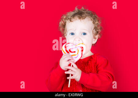 Beau Bébé curly girl eating a bonbons en forme de coeur sur fond rouge Banque D'Images