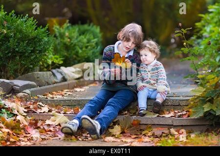 Son petit frère et petite sœur de parler et jouer avec les feuilles d'érable orange assis sur de vieux escaliers en pierre dans un parc d'automne Banque D'Images