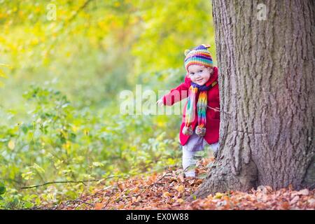 Sweet funny baby girl dans un manteau rouge et un chapeau et écharpe en tricot coloré se cacher derrière un gros arbre ancien dans un magnifique parc d'automne Banque D'Images