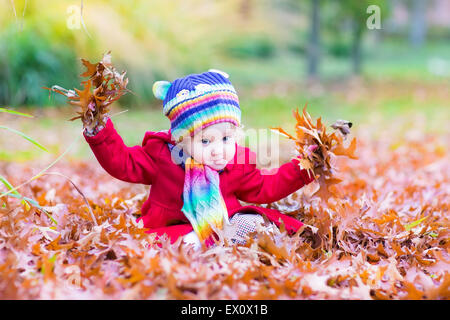 Adorable bébé fille avec un foulard et un chapeau coloré jouant avec feuilles rouges dans un parc d'automne Banque D'Images