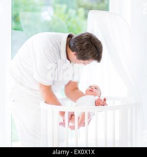 Young smiling père mettre son bébé nouveau-né dans une mangeoire ronde blanche à côté d'une grande fenêtre Banque D'Images