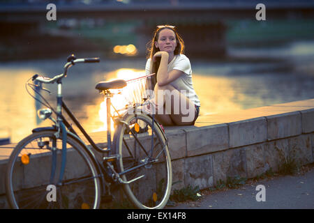 Adolescente avec le vélo, assis sur la promenade au coucher du soleil. Traitement croisé style du film. Banque D'Images