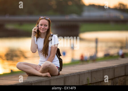 Jeune fille assise sur la promenade pendant le coucher du soleil et talking on mobile phone. Banque D'Images
