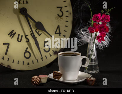 La vie encore d'une tasse de café sur une table avec une horloge dans l'arrière-plan ; un vase avec une fleur rouge, et quatre chocolats Banque D'Images