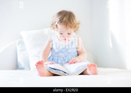 Funny Girl dans une robe bleue la lecture d'un livre sur un lit blanc dans une chambre à coucher de soleil Banque D'Images