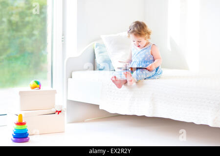 Adorable bébé fille lire un livre assis sur un lit blanc dans une grande fenêtre avec vue sur le jardin Banque D'Images