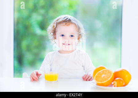 Funny curly girl drinking jus d'orange le matin dans une salle blanche avec une grande fenêtre Banque D'Images