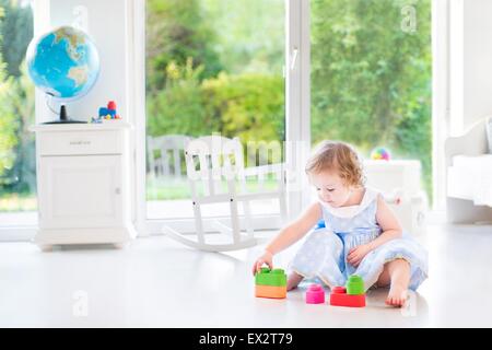 Petit bébé fille avec des cheveux bouclés portant une robe bleue jouant dans une chambre à coucher de soleil blanc avec une grande fenêtre avec vue sur jardin Banque D'Images