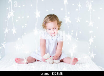 Magnifique petite fille jouant avec son ours en peluche sur un lit blanc entre les lumières de Noël Banque D'Images