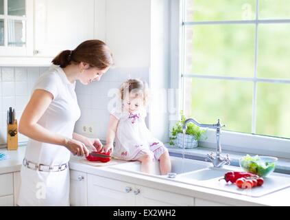 Mignon bébé fille avec des cheveux bouclés vêtue d'une robe blanche d'aider sa mère à faire cuire les légumes dans une belle cuisine ensoleillée Banque D'Images