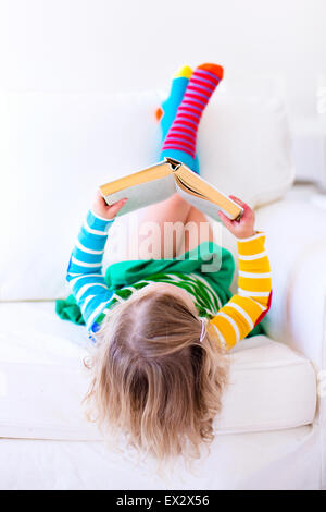 Petite fille lire un livre de vous détendre sur un canapé blanc. Lire des livres pour enfants à la maison ou à la garderie. L'apprentissage et les enfants à faire leurs devoirs Banque D'Images