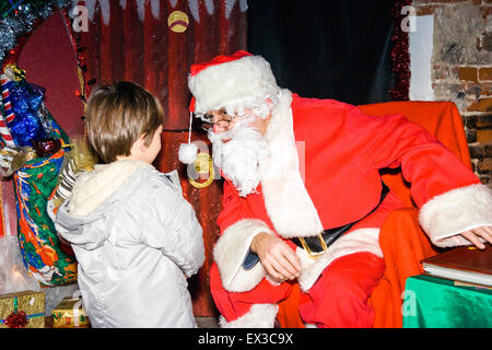 Boy in Santa's Grotto remis en question par le Père Noël. Caucasien enfant, 3-4 ans, porte manteau d'hiver. Santa entouré par des cadeaux. Banque D'Images