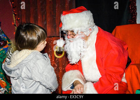 Boy in Santa's Grotto remis en question par le Père Noël. Caucasien enfant, 3-4 ans, porte manteau d'hiver. Santa entouré par des cadeaux. Banque D'Images
