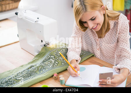 Happy Fashion designer holding smartphone et la rédaction de notes dans l'ordinateur portable Banque D'Images