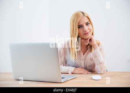 Happy young businesswoman sitting at the table with laptop and looking at camera sur fond gris Banque D'Images