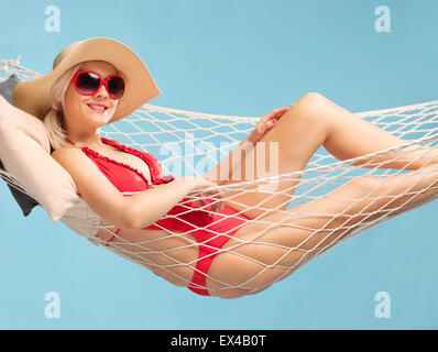 Belle femme dans un maillot rouge portant un chapeau élégant et couché dans un hamac sur fond bleu Banque D'Images