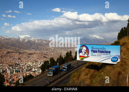 La Paz, Bolivie, le 6 juillet 2015. Un panneau d'accueillir le pape François à la Bolivie à côté de l'autoroute entre El Alto et La Paz, avec la ville de La Paz en dessous et la Cordillère Real montagnes (Mt Mururata, L, et le Mont Illimani, R) dans l'arrière-plan. Le pape François se rendra à La Paz le 8 juillet au cours de sa 3 journée à la Bolivie. Banque D'Images