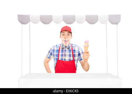 Les jeunes excités fournisseur avec un bonnet rouge et un tablier tenant un cornet de crème glacée derrière un stand de crème glacée isolé sur fond blanc Banque D'Images