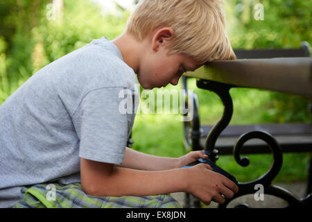 Enfant Garçon blond assis sur un banc de jardin à l'aide d'un mobile (cellulaire) smart phone, jeu de rôles, la messagerie texte, la concentration Banque D'Images