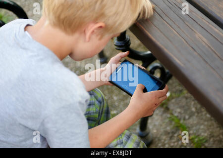 Enfant Garçon blond assis sur un banc de jardin à l'aide d'un mobile (cellulaire) smart phone, jeu de rôles, la messagerie texte, la concentration Banque D'Images