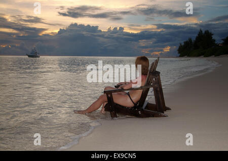 Une jeune femme est assise dans un fauteuil en bois du pont sur la rive de l'Océan Indien et admirer le coucher du soleil, Denis Island, Seychelles Banque D'Images