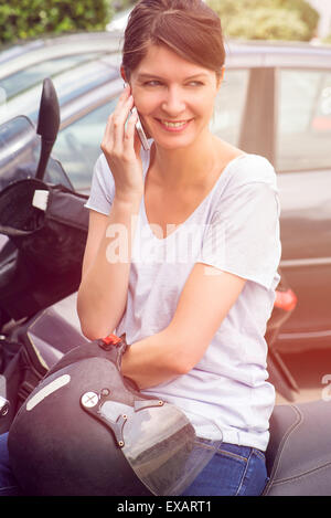 Femme assise sur moto garée, talking on cell phone Banque D'Images