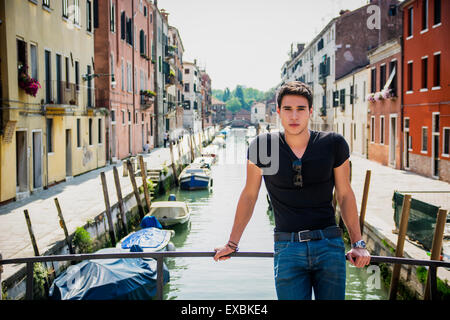 Portrait of Attractive Dark Haired Young Man Leaning Against Railing on pont pied plus petit canal à Venise, Italie Banque D'Images