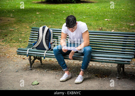 Jeune étudiant assis sur le banc avec les jambes croisés à côté de son sac à dos tout en étudiant ses leçons au sérieux. Banque D'Images