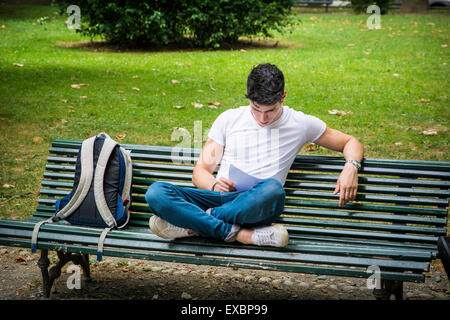 Jeune étudiant assis sur le banc avec les jambes croisés à côté de son sac à dos tout en étudiant ses leçons au sérieux. Banque D'Images