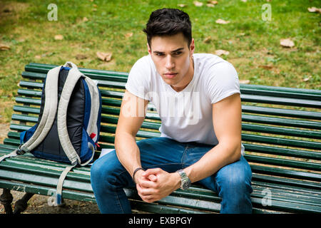 Jeune étudiant assis sur le banc dans un parc, à côté de son sac à dos, et tout en pensant à la suite au sérieux. Banque D'Images