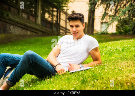 Young male student studying ses leçons en position couchée sur l'herbe au Parc de la ville, Smiling at Camera Banque D'Images