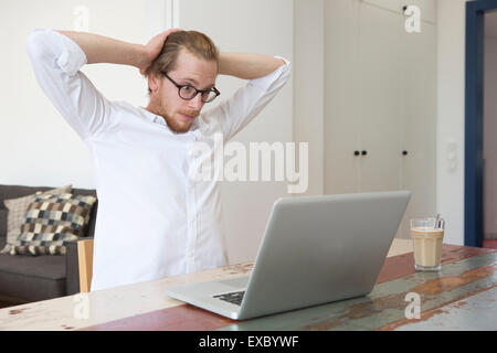 Red-haired young man sitting at table avec son ordinateur portable et à la recherche a souligné et excité Banque D'Images