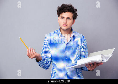 Jeune homme sérieux tenant le crayon et dossier avec des fichiers sur fond gris. Looking at camera Banque D'Images