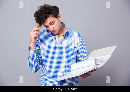 Pensive young man holding pencil et dossier avec des fichiers sur fond gris. Looking at camera Banque D'Images