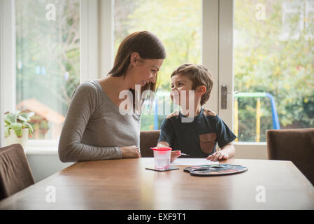 Boy smiling à mère en tant qu'il peint sur papier à table dans la salle de séjour Banque D'Images