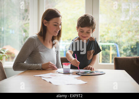 Boy smiling with mother comme il peint sur papier à table dans la salle de séjour Banque D'Images