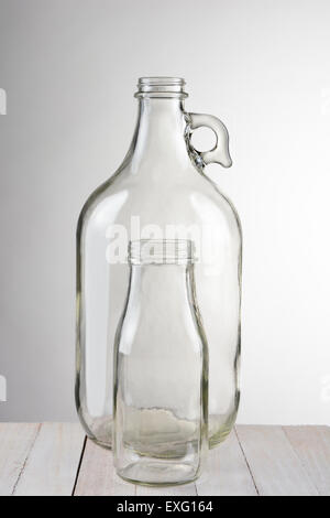 Deux bouteilles en verre vide, une petite bouteille en face d'un plus grand récipient. Format vertical contre une lumière sur fond gris foncé. Banque D'Images