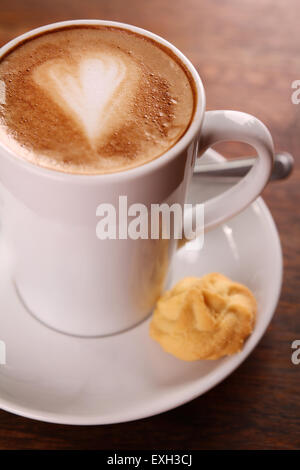 De délicieux cappuccino aromatique dans une tasse de porcelaine blanche avec un biscuit sur le côté sur une table en bois. Banque D'Images