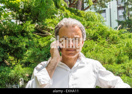 Bel homme d'âge moyen avec des cheveux poivre sel habillé avec une chemise blanche est talking on mobile phone in city park : il montre un regard rassurant Banque D'Images