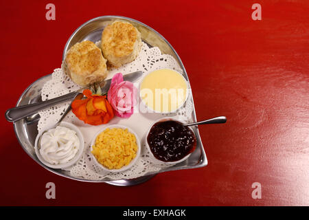 Délicieux scones beurre sur un plateau en forme de coeur argent servi avec du fromage et de la confiture de roses crème conserve sur une table en bois rouge. Banque D'Images