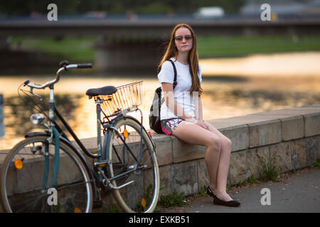 Charmante teen girl avec le vélo assis sur la berge de la rivière au coucher du soleil. Traitement croisé style photo. Banque D'Images