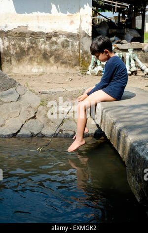 Petit garçon assis tout seul sur le petit pont de pierre sur la pêche à l'eau avec un morceau de ficelle dans un village proche de java solo Banque D'Images
