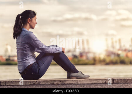 Vue arrière d'une jeune femme face à la rivière tout en restant assis sur un long banc en béton et serrant ses jambes Banque D'Images