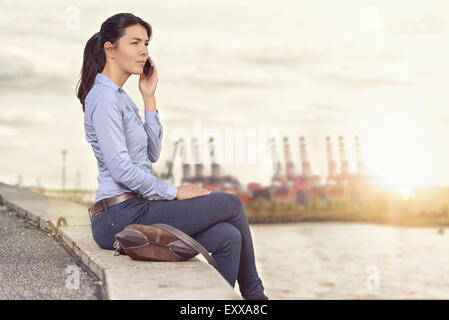 Jolie femme élégante de parler sur son téléphone portable pendant qu'elle se trouve sur un mur donnant sur une rivière et ville éloignée, avec vue latérale Banque D'Images