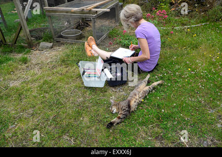 Artiste féminin assis dehors à côté d'un chat qui s'étend des croquis canards dans une cage en été dans les régions rurales du pays de Galles UK KATHY DEWITT Banque D'Images